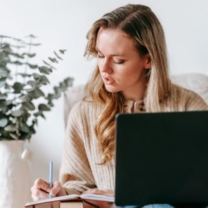 Woman in cozy attire working on a laptop and writing notes in a home setting.