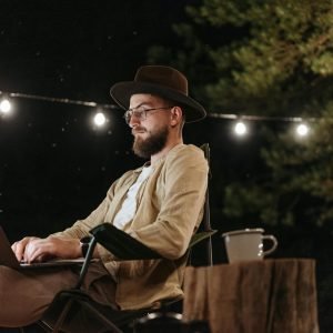 A man sits outdoors using a laptop at night, surrounded by string lights.